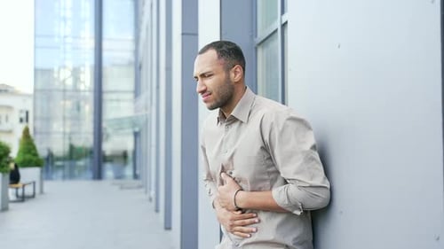 Adult man feels stomach pain while standing on the street near an office building. A sad male of mix