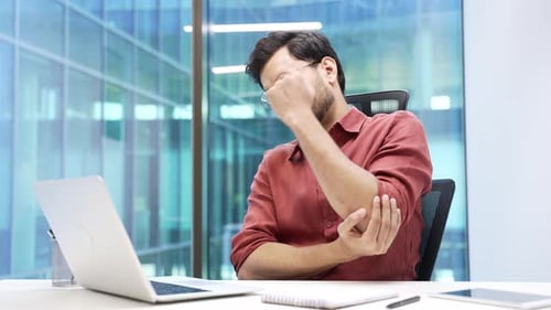 Businessman suffering from elbow pain while working on a laptop sitting at desk in a business office
