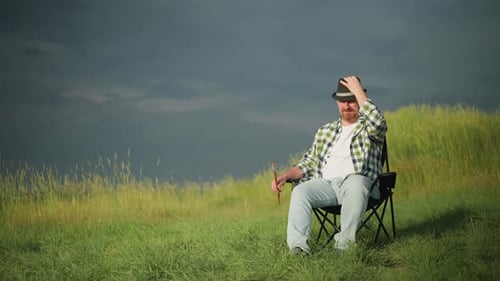 Man Adjusting Hat with Paintbrush in Hand Sitting in Field Under Dark Sky