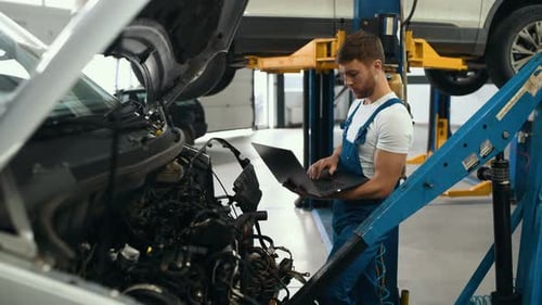 Handsome mechanic in uniform is using a laptop while repairing car in auto service