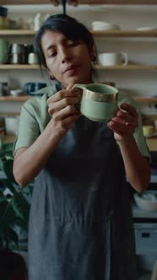 Craftswoman in Apron Holding Ceramic Mug and Examining It in Potters Studio