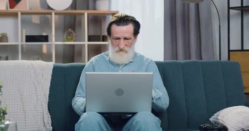 Likable senior man working on laptop computer sitting on the sofa in cozy living-room