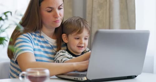 Mother Working on Laptop From Home Remotely with Little Son in Her Arms