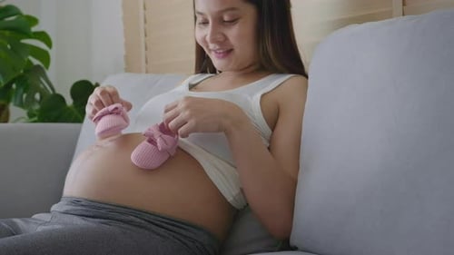 Pregnant Woman Holding Pink Baby Shoes on Couch