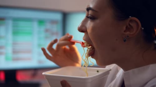 Closeup View of Woman Eating Instant Noodles Sitting in Office Against Computer Screens Background