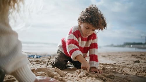 Cute curly boy building sand figures at the ocean shore with mother in autumn sunlight