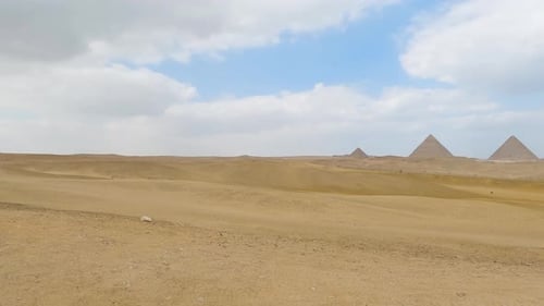 Pan across sand desert dunes to view of three pyramids off in the distance