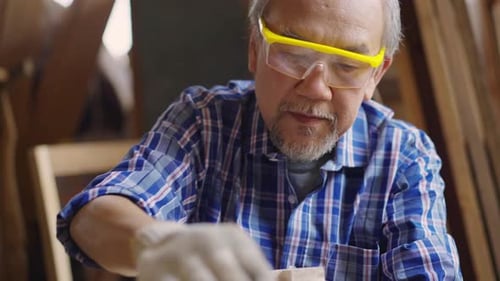 An elderly Asian male carpenter sanding a wooden piece of furniture in a carpentry workshop