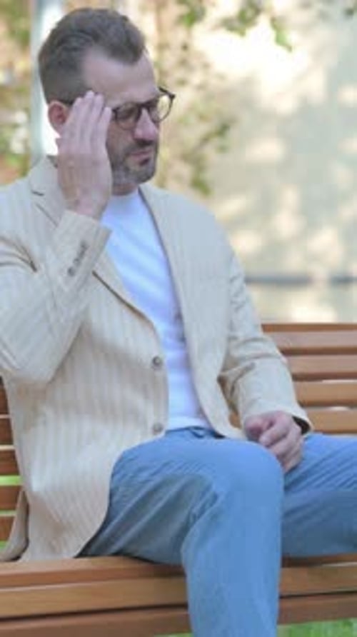 Bearded Man Sitting on Bench Rubs Temples