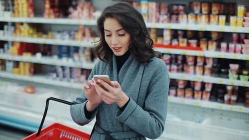 Brunette Young Woman Walk Checking to Do List on Phone in Supermarket Smiling Thinking Shopping