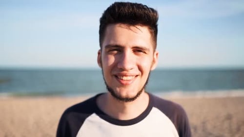 Portrait of Young Man Looking and Smiling at Camera Standing on the Beach with a Sea on Background