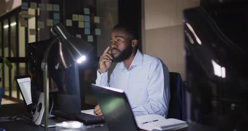 Video of african american businessman sitting at desk using computer at night in office
