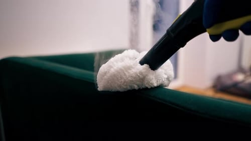 Close Up in an Apartment Cleaner in a Yellow Apron Does a Steam Cleaning of a Green Sofa with