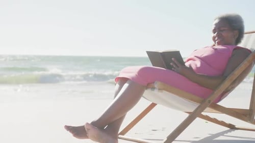 Happy senior african american woman sitting on deck chair and reading book at beach, in slow motion