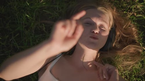 Weiße Frau im Gras liegend mit Kopfhörern, sonniger Nachmittag, sanfte Brise, Fingertanz, Augen geschlossen