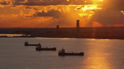 Aerial drone view of multiple oil tankers anchored near an industrial port at sunset with cranes and