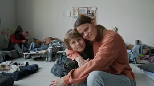 Portrait of Young Female Refugee Hugging and Kissing Son in Shelter