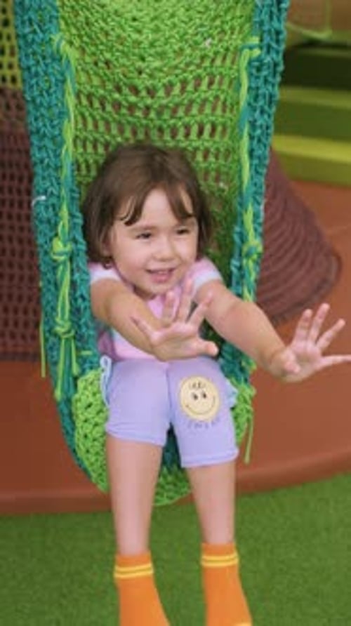 Garota sentada em um balanço de crochê no parquinho coberto, estendendo as mãos alegremente.