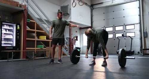 Woman deadlifts barbell with coach in indoor gym