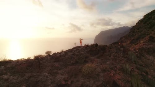Adult girl stands with arms outstretched on a rocky hill, gazing at the ocean and cliffs, under a so