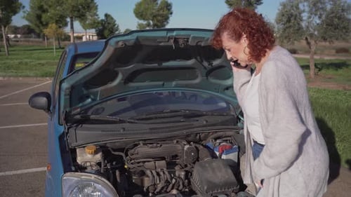 Senior woman standing by broken-down car with hood open calling for roadside assistance