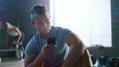 Handsome and Masculine Athletic Young Man is Using a Smartphone while Sitting on a Floor in a Loft