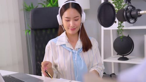 Asian podcaster in headphones using microphone broadcasting in studio.