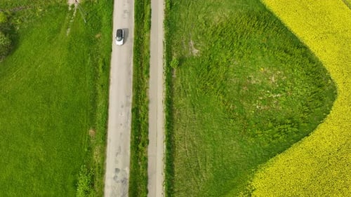 Aerial view of a rural road with a white car driving along it, bordered by green fields and a yellow