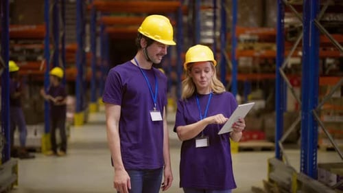Two warehouse workers wearing safety helmets and badges are using a digital tablet, discussing logis