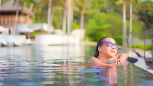 Beautiful young Asian woman wearing sunglasses resting at side of infinity swimming pool of luxury t