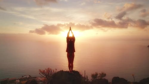 Person stands with arms raised towards the sunset on a rocky cliff, surrounded by the sea and a beau