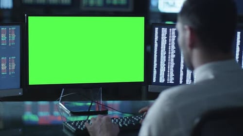 Man is working on a computer with mock-up green screen in a dark office filled with displays.