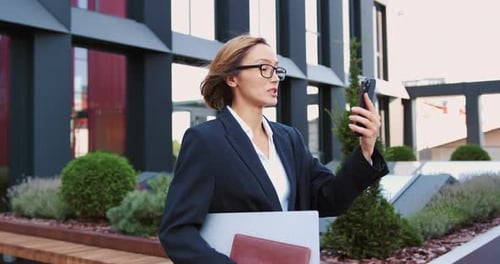 Business woman using smartphone outdoors standing on city street, looking to mobile phone screen, en