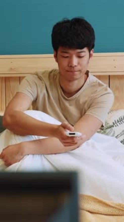 A Young Man is Comfortably Lying in Bed Casually Holding a Remote Control in His Hand