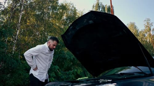 a young bearded man stands near a car with an open hood and tries to check a breakdown