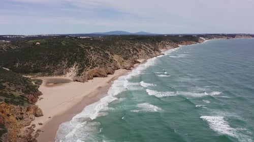 Aerial of wild stormy ocean waves under cloudy sky and sandy beach, with cliffs along coastline