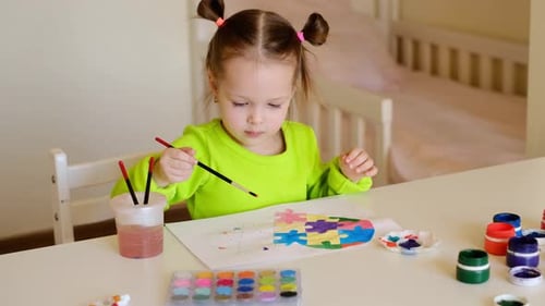 The girl dips paintbrush in watercolor paints on palette and paints drawing with heart-shaped puzzle