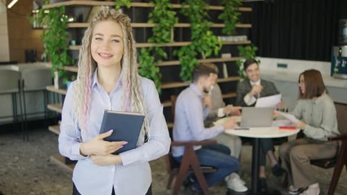 Young Woman with Tablet Smiling Confidently in a Modern Office Setting While a Group of Colleagues