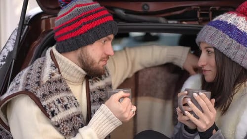 Side View Portrait of Happy Young Couple Drinking Hot Tea Sitting in Car Trunk on Winter Day