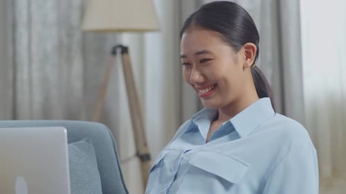 Close Up Of Smiling Asian Woman Typing On A Laptop Computer While Lying On Sofa In The Living Room