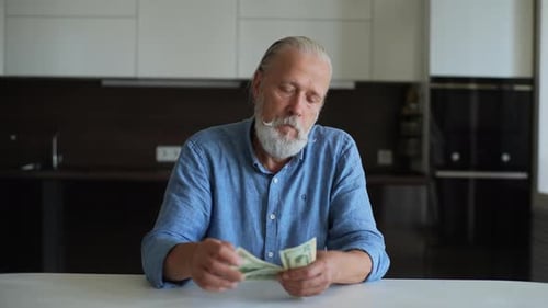 Portrait of Sad Angry Senior Pensioner Male Counting Dollars Looking Upset Sitting at Home Desk