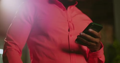 Man of African descent in coral shirt holding smart phone outdoors at night, cropped view focused