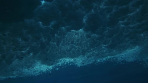 Breaking wave crashes as bubbles spread across ocean surface twirling in vortex, underwater angle