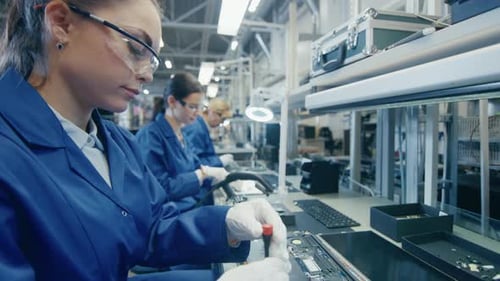 Une travailleuse d'une usine d'électronique en blouse de travail bleue et lunettes de protection assemble un ordinateur portable Mo
