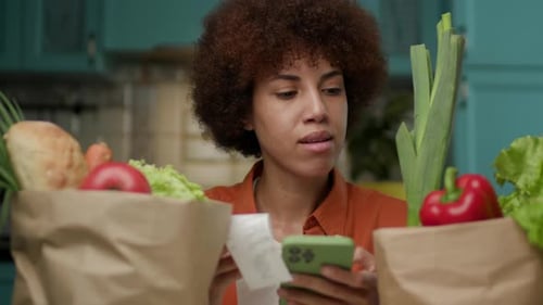 Mulher verificando recibo de mercearia Mulher negra olhando para sacolas cheias de alimentos frescos e celular
