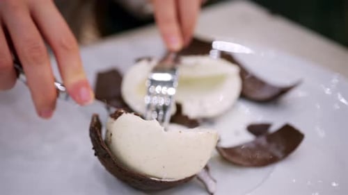 Close up of a dessert shaped like a coconut being opened with a fork, revealing a creamy white inter