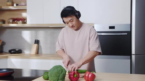 Close Up Young Asian Man Cooking at Kitchen Wearing Headphones Cutting Fresh Cucumber on Wooden
