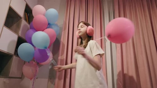 Jugendliches Mädchen, das aufgeregt Luftballons in einer gemütlichen Innenumgebung mit rosa Vorhängen und Ohrenschützern hält