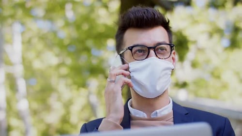 Businessman in city park works on laptop and smartphone wearing face mask