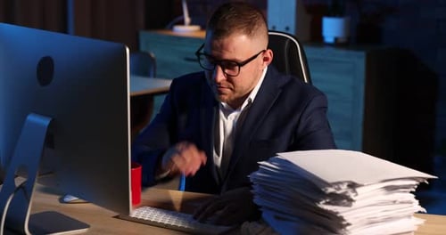 Bourreau de travail. Un homme submergé et des lunettes près d'une pile de documents sur un bureau en bois à l'intérieur la nuit.
